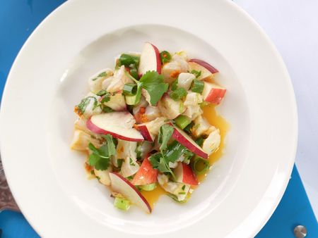Draufsicht auf einen weißen Teller mit Kabeljau-Ceviche, garniert mit Pfirsichspalten, Koriander, Frühlingszwiebeln und einem orangefarbenen Sanddorn-Dressing, angerichtet auf einem blauen Schneidebrett.