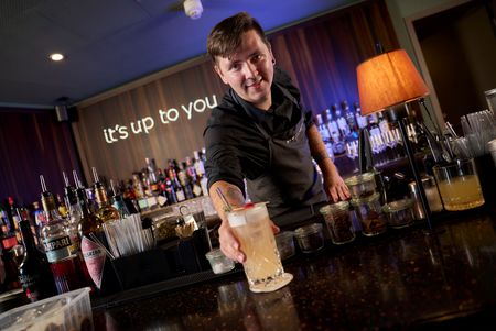 Barchef Gleb Filippov in schwarzer Arbeitskleidung reicht einen Cocktail im Highball-Glas über einen dunklen Bartresen. Im Hintergrund befinden sich Spirituosenflaschen und der beleuchtete Schriftzug „it's up to you".