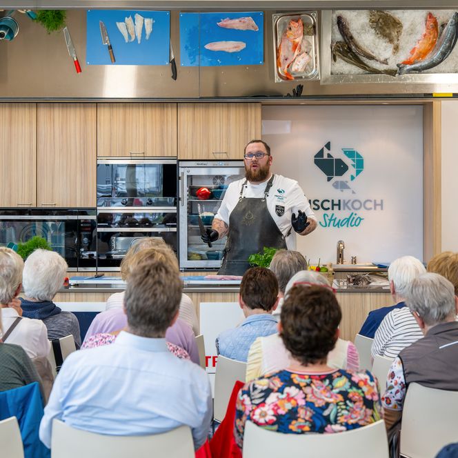 Koch erklärt vor Publikum die Zubereitung von Fischgerichten in moderner Studioküche in Bremerhaven.