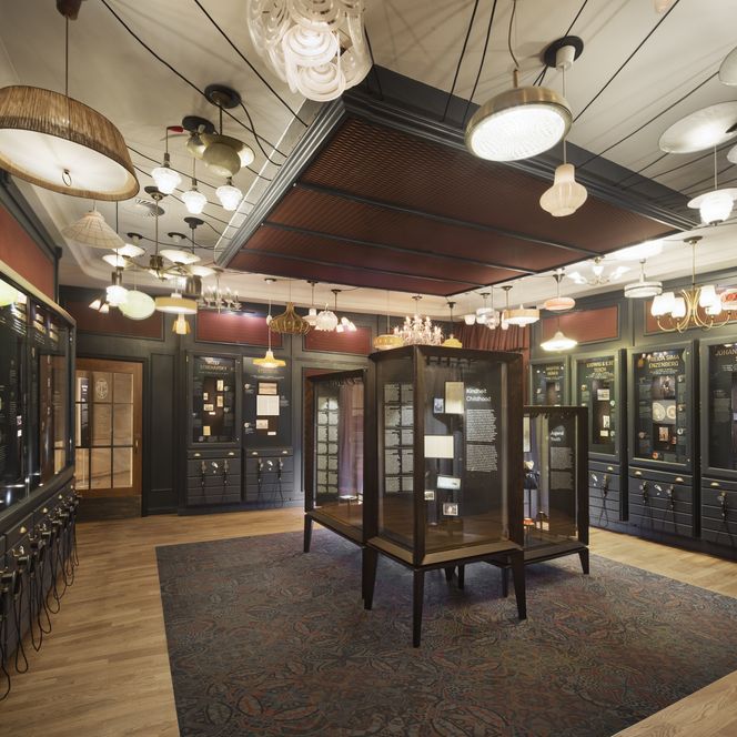 Ausstellungsraum mit Vitrinen, historischen Dokumenten und vielfältigen Lampen an der Decke