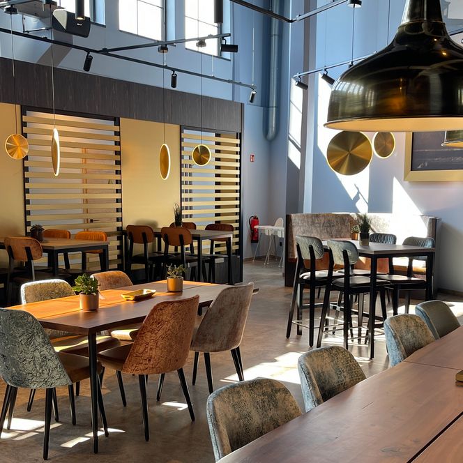 Helles Restaurant mit modernen Holztischen, Polsterstühlen und großen goldfarbenen Lampen.