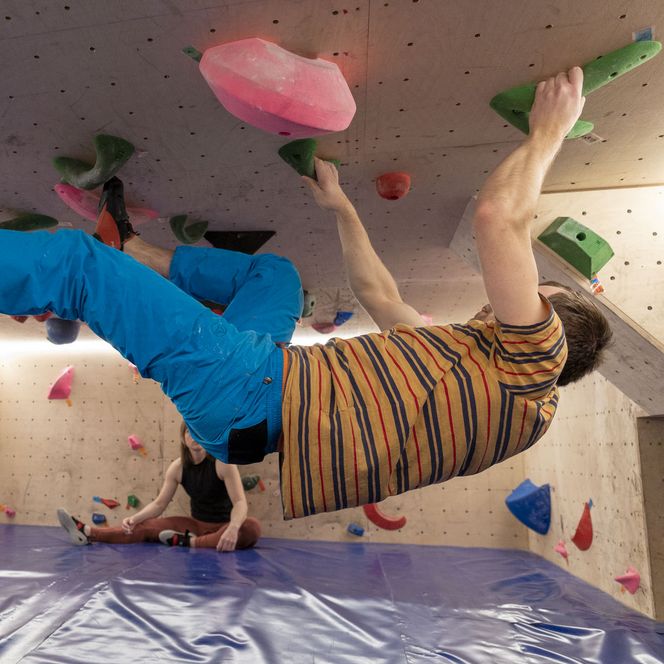 Person beim Bouldern an überhängender Kletterwand mit bunten Griffen in der Halle.