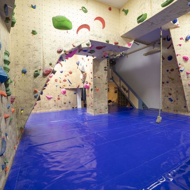 Innenraum einer Kletterhalle mit bunten Griffen, blauen Matten und Seil zum Training an der Wand.