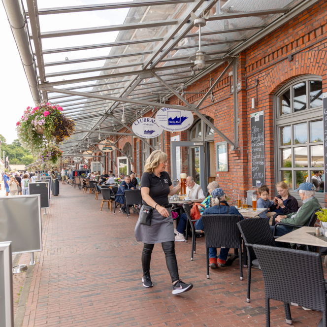 Menschen sitzen vor Backsteinfassade in Bremerhaven beim Essen unter überdachter Promenade.