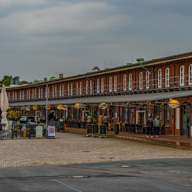 Backsteinbau mit überdachter Promenade, Restaurants und Spaziergängern im Schaufenster Fischereihafen.