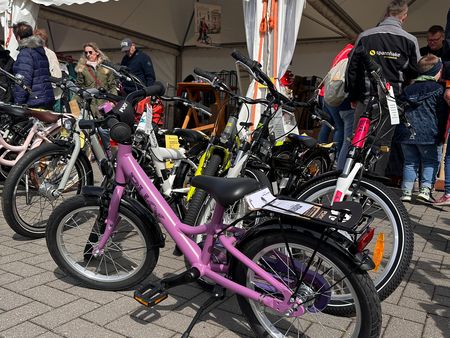 Mehrere Kinderfahrräder, darunter ein lilafarbenes im Vordergrund, stehen auf einem Pflasterplatz unter weißen Messezeltdächern. Im Hintergrund besichtigen Besucher das Ausstellungsangebot.