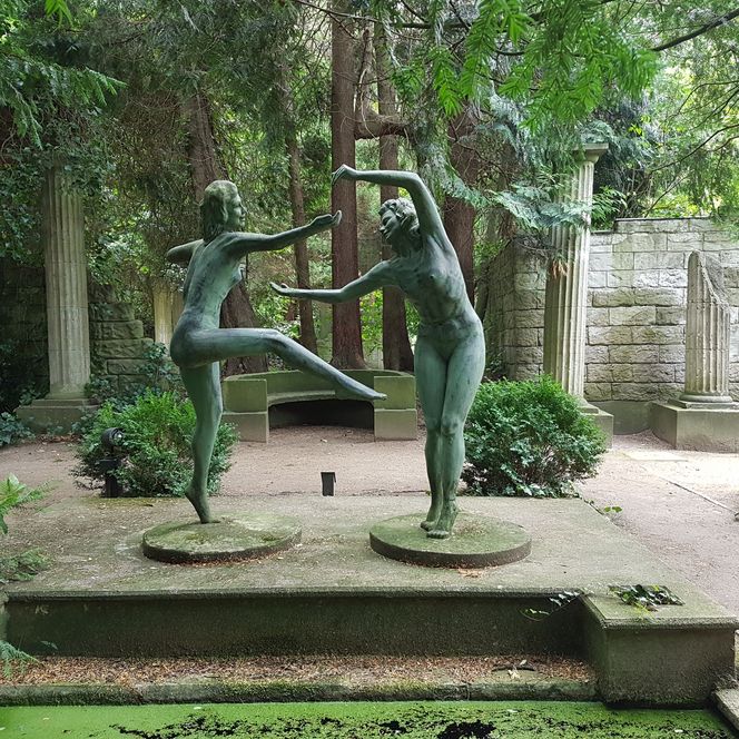 Bronzefiguren tanzender Frauen vor alten Steinsäulen, umgeben von dichtem Grün in Thieles Garten.