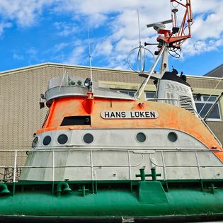Nahaufnahme des Aufbaus des Museumsschiffs „Hans Lüken" mit orangefarbenem Brückenhaus, weißem Rumpfbereich mit Bullaugen, grünem Unterdeck und technischen Aufbauten vor einem teils bewölkten blauen Himmel und einem Backsteingebäude im Hintergrund.