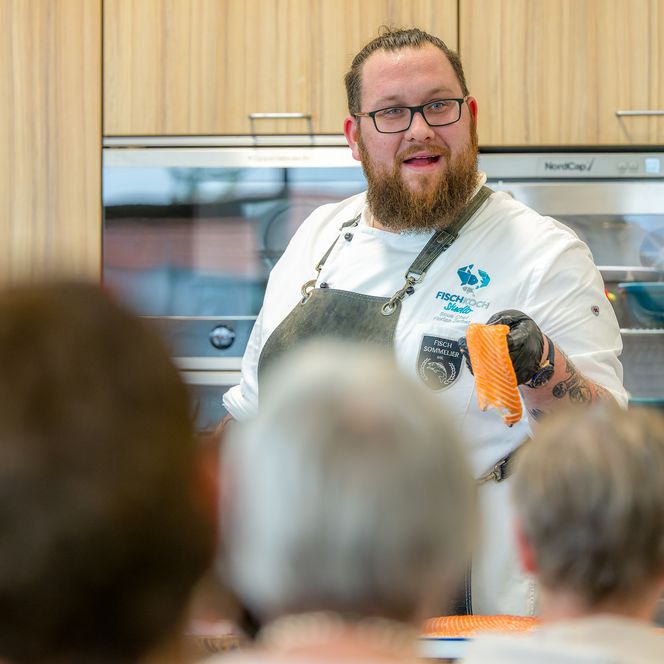 Ein Koch demonstriert vor Publikum die Zubereitung eines Stücks Lachs in einer modernen Küche.