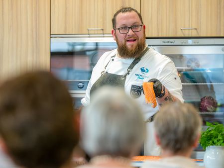 Ein Koch demonstriert vor Publikum die Zubereitung eines Stücks Lachs in einer modernen Küche.