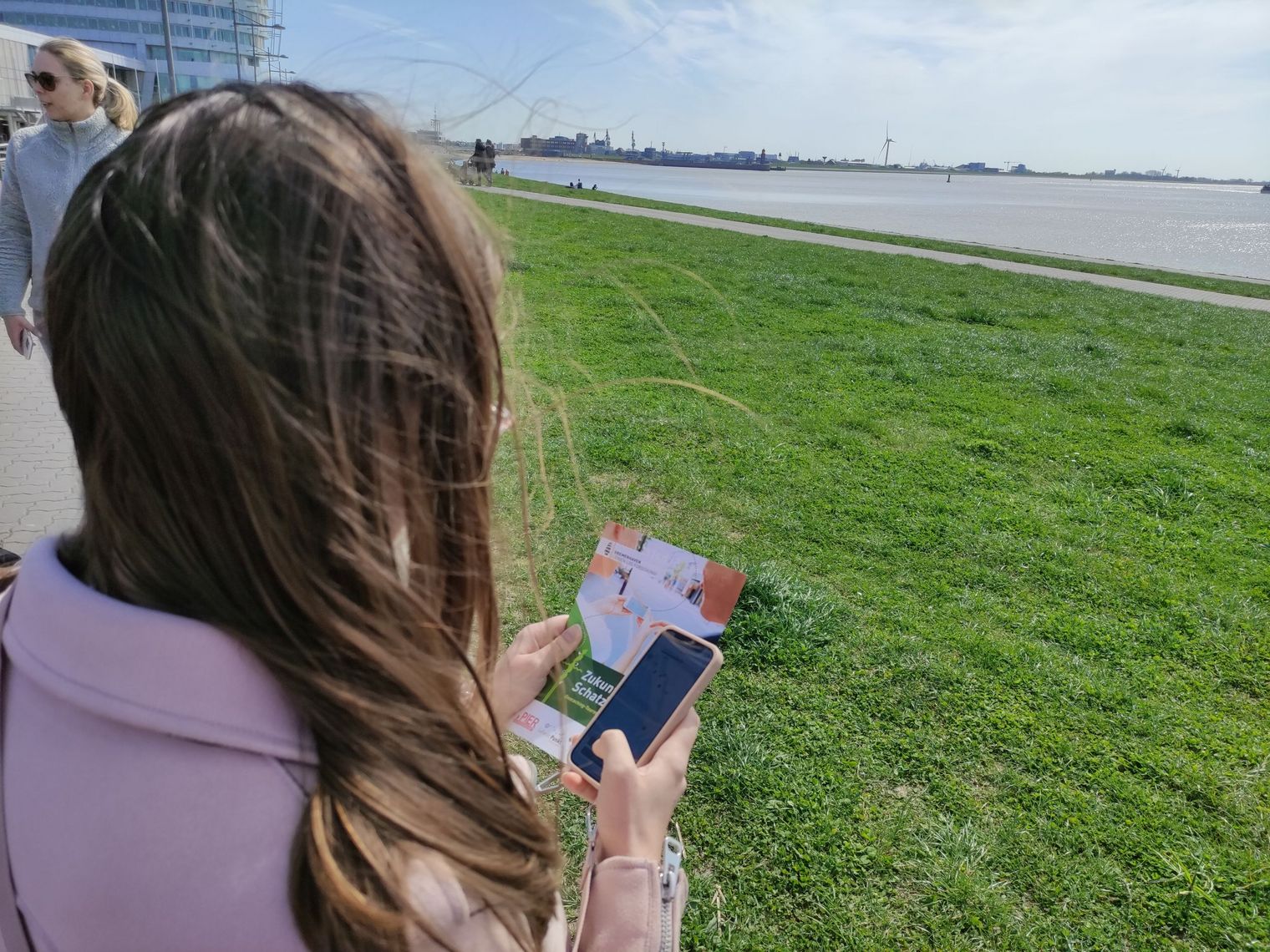 Eine Frau steht am Deich und führt über eine App am Handy die Zukunftsschatzsuche in Bremerhaven durch.