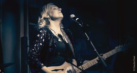 Vanesa Harbeck steht mit geschlossenen Augen singend vor einem Mikrofon auf einer Bühne und hält eine E-Gitarre. Sie trägt eine schwarze Spitzenjacke und Ketten. Das Bild ist in Blautönen gehalten.