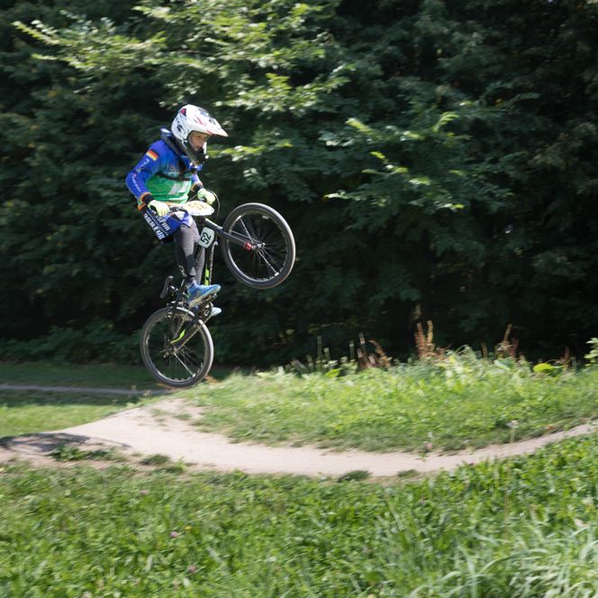 Junge Person springt mit BMX-Fahrrad auf einer Dirt-Strecke vor grünem Hintergrund.