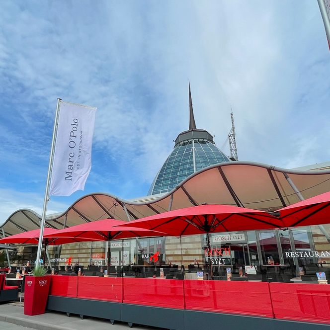 Außenbereich eines Restaurants mit roten Sonnenschirmen vor dem modernen Shopping-Center Gebäude.