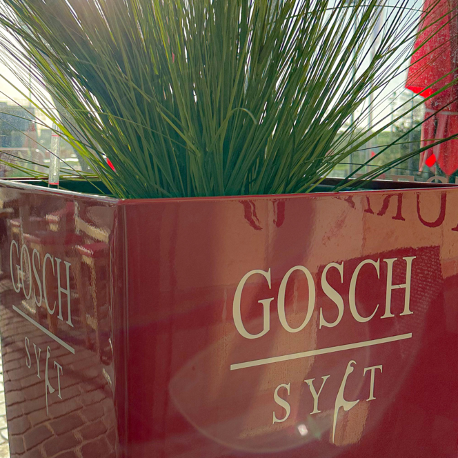 Roter Gosch Sylt Pflanzkübel mit grünem Gras auf der Außenterrasse am Hafen.