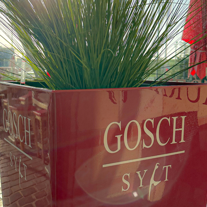 Roter Gosch Sylt Pflanzkübel mit grünem Gras auf der Außenterrasse am Hafen.