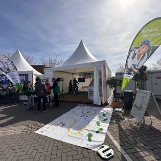 Messestand der Deutschen Verkehrswacht mit weißem Pagodenzelt, grünen Beachflags mit Aufschrift „FahrRad… aber sicher!", einem Parcours-Spielfeld auf dem Boden sowie Besuchern und Standpersonal im Freien bei sonnigem Wetter.