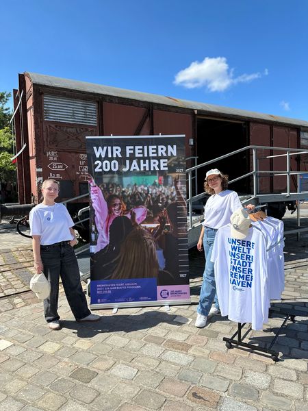 Zwei Frauen vor einem Banner mit der Aufschrift "Wir feiern 200 Jahre"