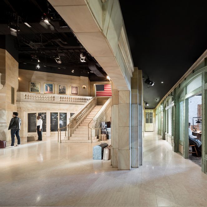 Weitläufige Ausstellungshalle mit Marmortreppen, Reisenden, Gepäckstücken und US-Flagge an der Wand.