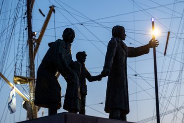 Dunkle Bronzestatue einer Auswandererfamilie vor beleuchteten Schiffsmasten in der Dämmerung.
