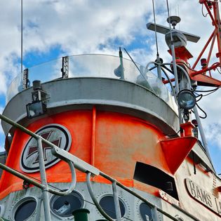 Detailansicht des orangefarbenen Brückenturms des Seenotrettungsschiffs "Hans Lüken" mit Navigationsantennen, Scheinwerfer, rundem Steuerstand und dem Schiffsnamen am Rumpf, vor bewölktem Himmel.