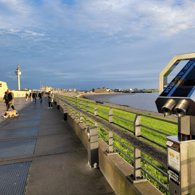 Aussichtsfernrohr an der belebten Deichpromenade in Bremerhaven. Menschen spazieren am Wasser.