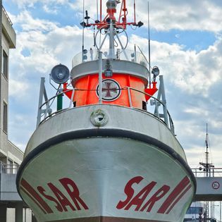 Frontansicht des orangeweißen Seenotrettungskreuzers „Hans Lüken" mit der Aufschrift „SAR" am Bug, aufgestellt an Land im Hafen von Bremerhaven vor bewölktem Himmel.