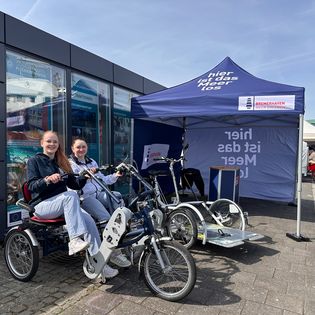 Zwei lächelnde junge Frauen sitzen auf einem blauen Duorad vor dem Messestand von Erlebnis Bremerhaven mit dunkelblauem Pavillon und der Aufschrift „hier ist das Meer los".