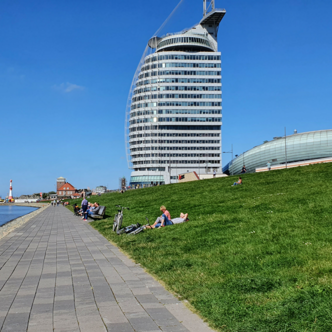 Deichpromenade Bremerhaven am Wasser. Menschen auf dem grünen Deich vor segelförmigem Hotel.