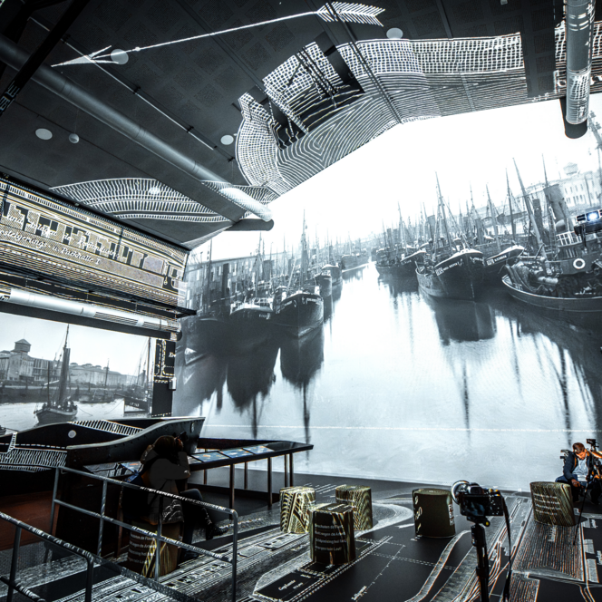 Innenraum mit großflächiger Projektion historischer Fischkutter im Hafen, Besucher fotografieren die Szene.
