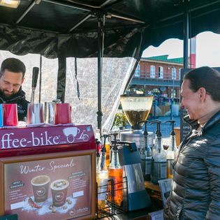 Barista an einem Coffee-Bike-Stand bereitet Kaffee zu, eine lächelnde Frau steht als Kundin davor. Auf der Theke sind Sirupflaschen, eine Kaffeemühle und ein Schild mit Winter Specials zu sehen.