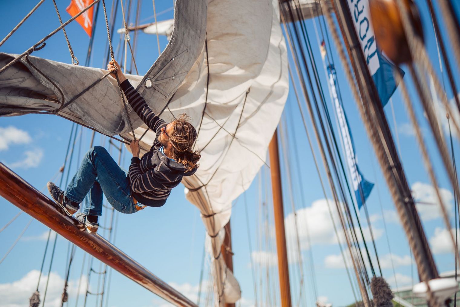 Mann hängt in den Rahen eines Segelschiffes und hisst ein Segel