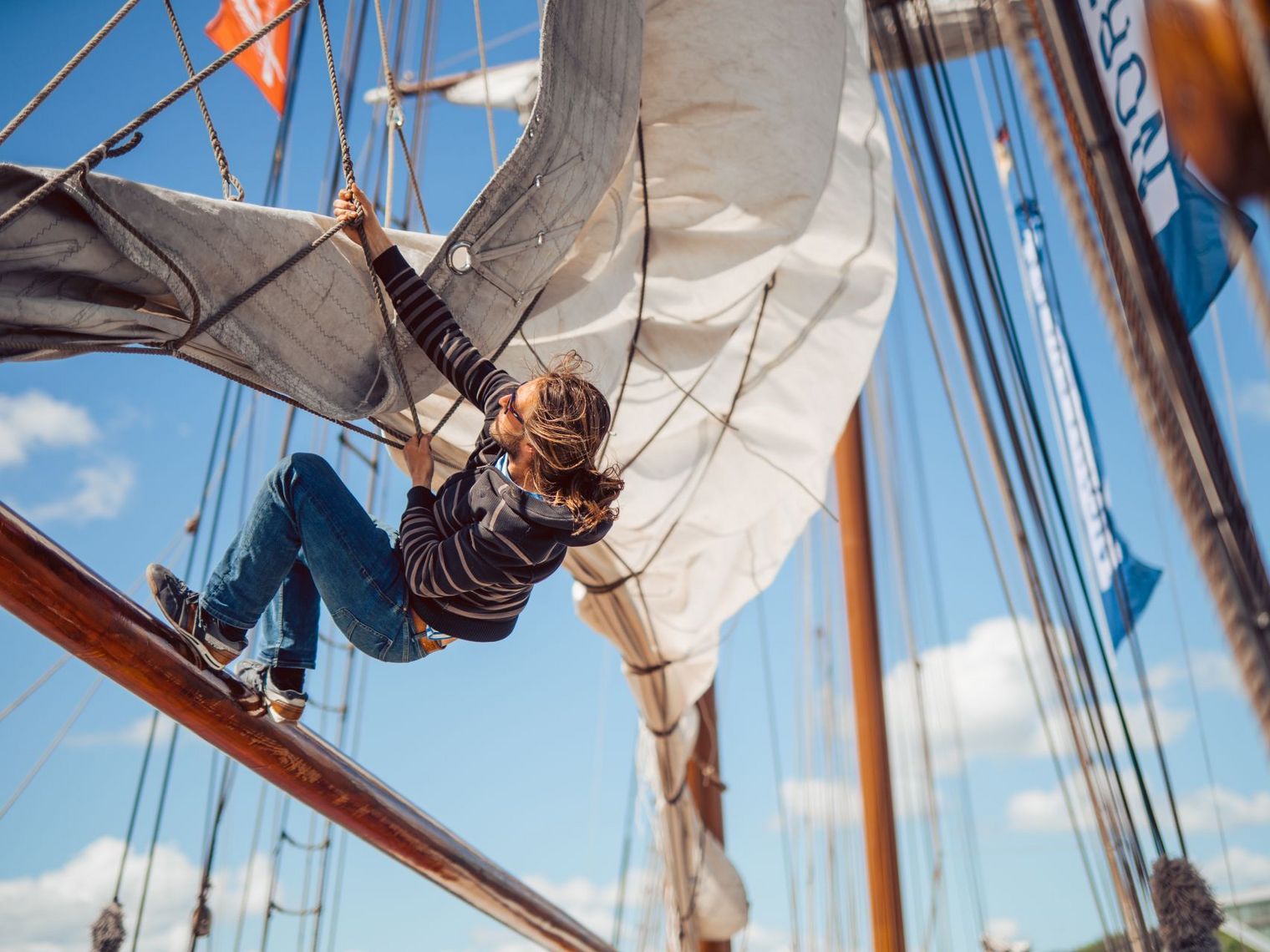 Mann hängt in den Rahen eines Segelschiffes und hisst ein Segel