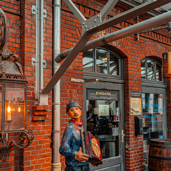 Backsteinfassade mit großer Laterne, Holzfigur eines Akkordeonspielers und Eingang zu Fiedlers Fischmarkt.