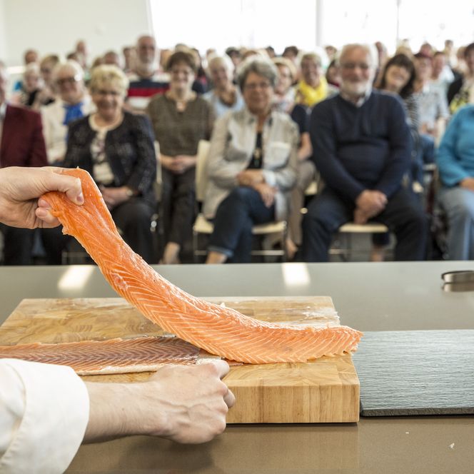 Koch präsentiert frisches Lachsfilet vor interessiertem Publikum in moderner Showküche.