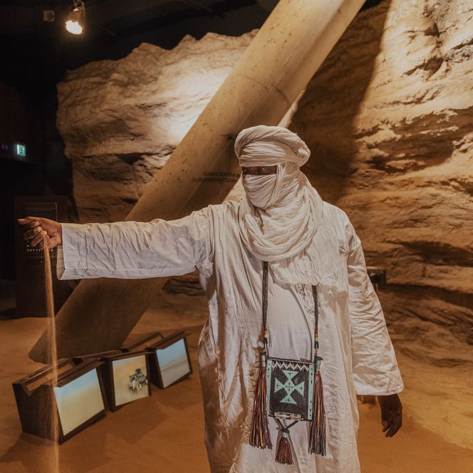 Mann in weißem Tuchgewand steht in sandiger Ausstellung und lässt Sand durch die Hand rieseln.