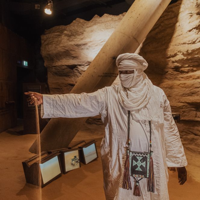 Mann in weißem Tuchgewand steht in sandiger Ausstellung und lässt Sand durch die Hand rieseln.