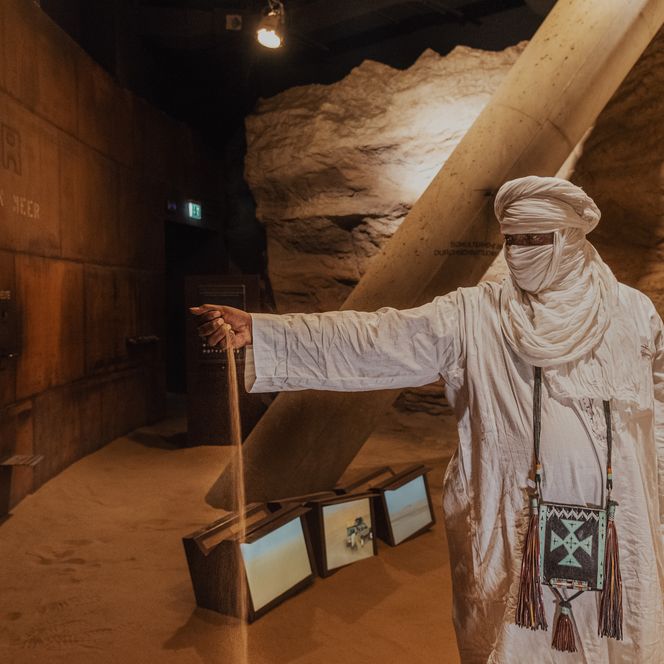 Mann in weißem Tuchgewand steht in sandiger Ausstellung und lässt Sand durch die Hand rieseln.