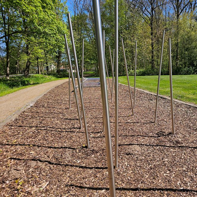 Fitnessstation mit senkrechten Metallstangen auf Rindenmulch in einem grünen Park.