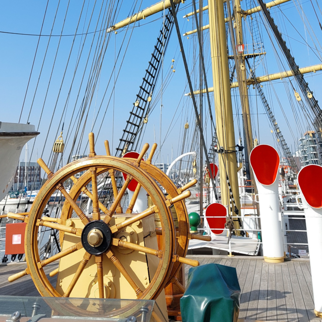 Holzsteuerrad auf dem Deck des Schulschiffs Deutschland vor gelben Masten und Tauwerk im Hafen.