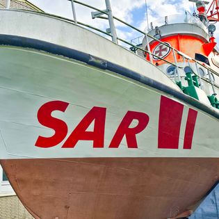 Bug des Seenotrettungskreuzers "Hans Lüken" mit großem rotem SAR-Schriftzug auf weißem Schiffsrumpf, aufgebockt vor einem Gebäude in Bremerhaven.