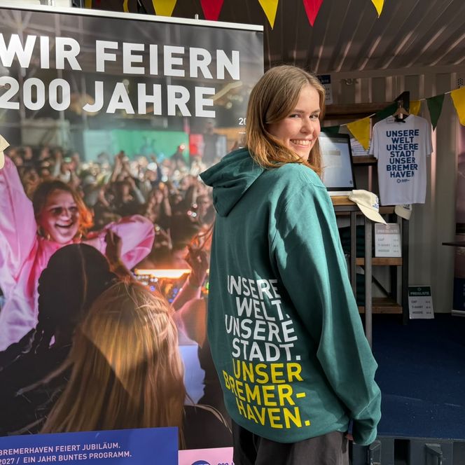 Junge Frau trägt grünen Kapuzenpullover mit Jubiläums-Slogan an einem Merchandise-Stand für das 200-jährige Bestehen Bremerhavens.