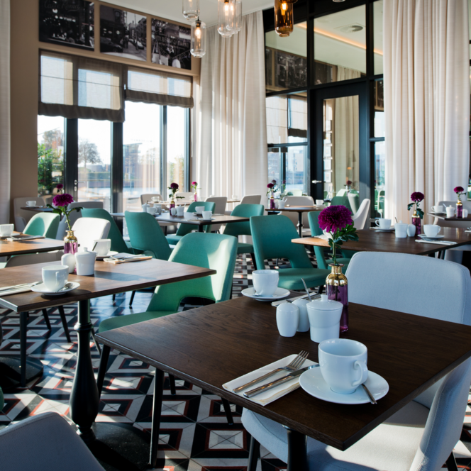 Helles Restaurant mit gedeckten Holztischen, türkisen Stühlen und geometrischem Fliesenboden.