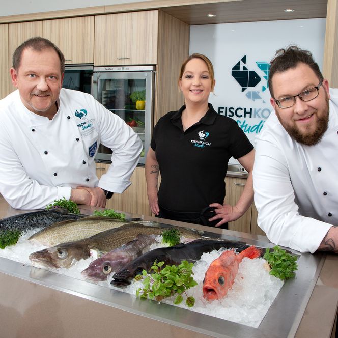 Drei Personen in Kochkleidung stehen in einer modernen Küche hinter frischem Fisch auf Eis.