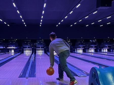 Person beim Bowling in beleuchteter Halle, wirft orange Kugel auf leuchtende Pins unter blauem Licht.