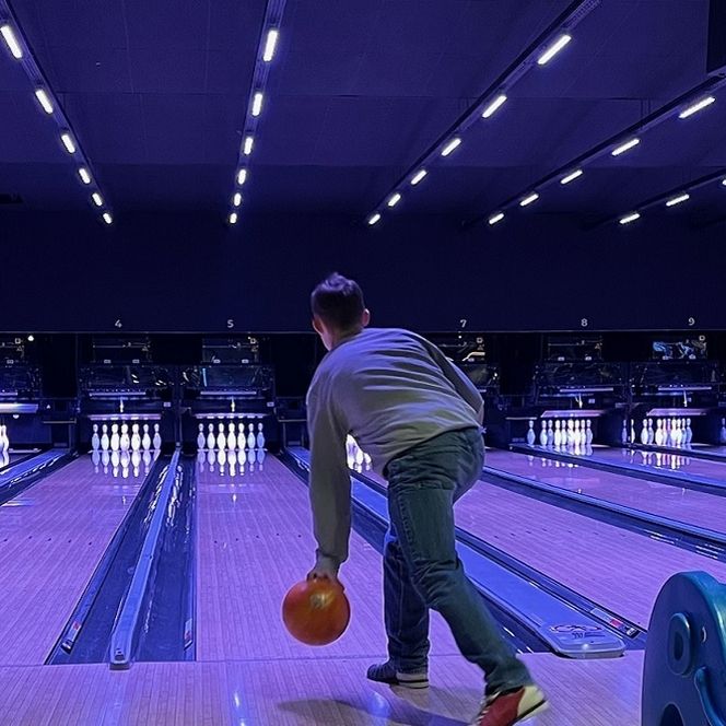 Person beim Bowling in beleuchteter Halle, wirft orange Kugel auf leuchtende Pins unter blauem Licht.