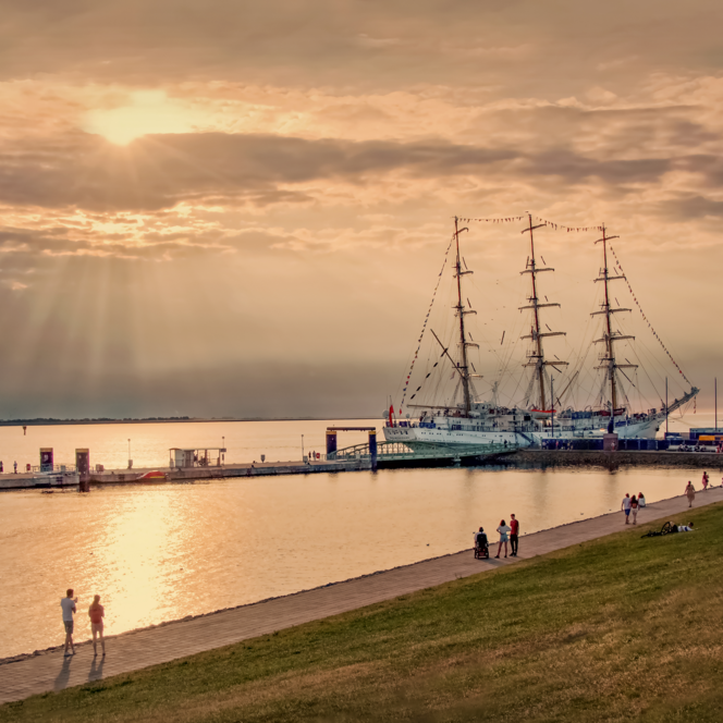 Spaziergänger auf der Deichpromenade im Gegenlicht. Am Pier liegt ein großes Segelschiff.