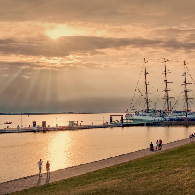 Spaziergänger auf der Deichpromenade im Gegenlicht. Am Pier liegt ein großes Segelschiff.