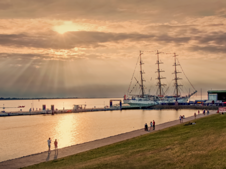 Spaziergänger auf der Deichpromenade im Gegenlicht. Am Pier liegt ein großes Segelschiff.